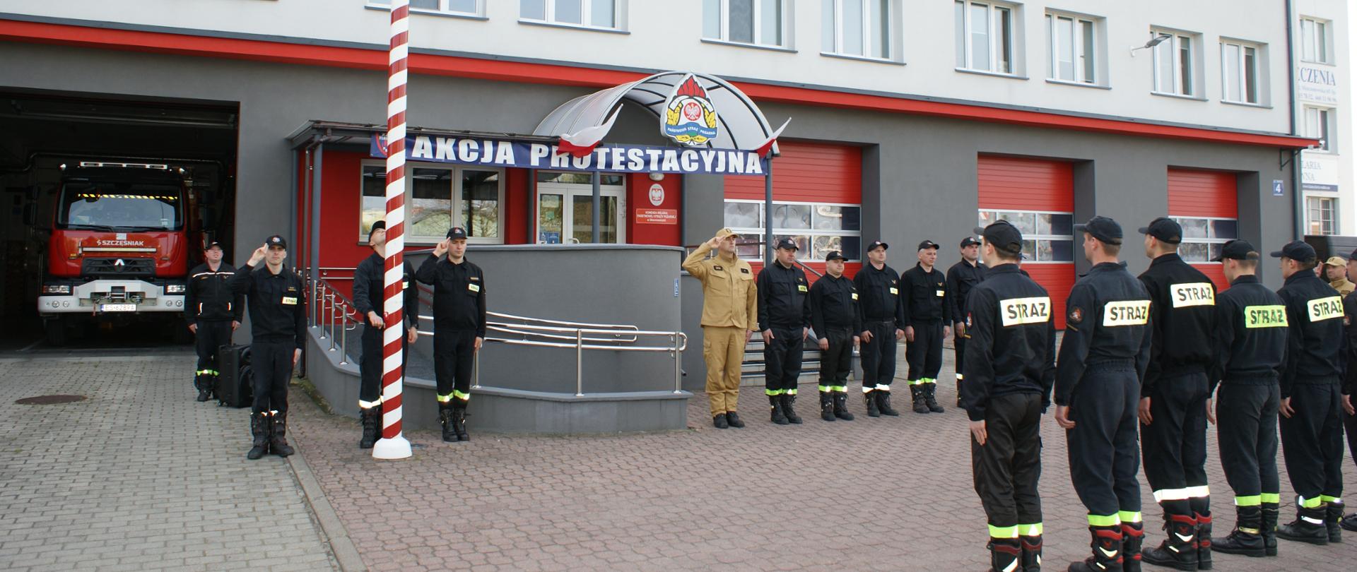 Na zdjęciu strażacy w ubraniach koloru piaskowego i czarnego stoją naprzeciw siebie przed budynkiem, w tle zaparkowane pojazdy oraz maszt flagowy.