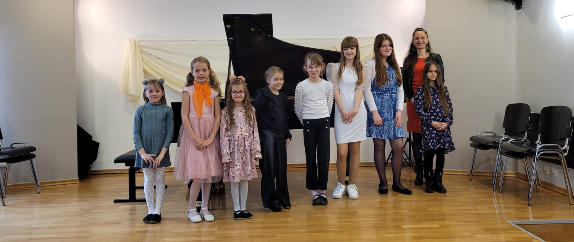 Na zdjęciu grupa uczniów- same dziewczyny wraz z nauczycielką- blondynką w czerwonej sukience i czarnej marynarce. W tle otwarty fortepian. Wszyscy stoją na scenie.