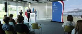 Minister funduszy i polityki regionalnej Katarzyna Pełczyńska-Nałęcz stoi przy mównicy, po jej prawej stronie stoi wiceminister Jan Szyszko i tłumaczka języka migowego, za ich plecami flagi Polski i UE, a po lewej ekran ze zdjęciem lotniska we Wrocławiu