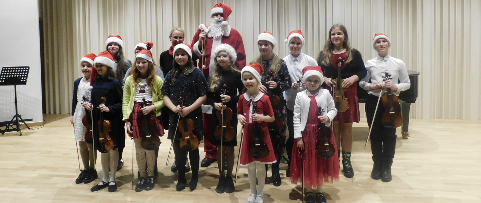 Na zdjęciu grupa uczniów, którzy trzymają skrzypce i mają założone czerwone czapeczki mikołajkowe. Za nimi stoi w czerwonym stroju Święty Mikołaj.