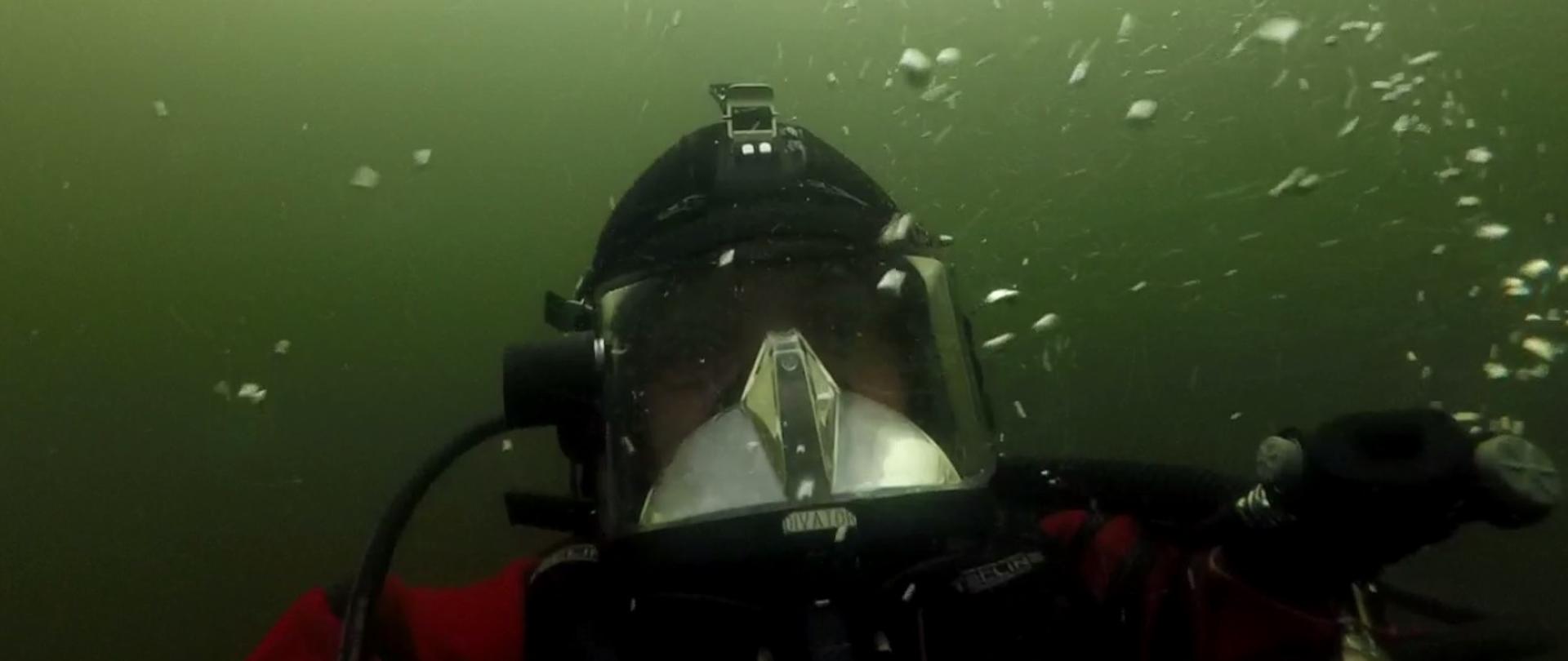 Zdjęcie przedstawia twarz nurka pod wodą. Strażak ma założoną maskę do nurkowania. Dookoła widoczne są pęcherzyki powietrza.