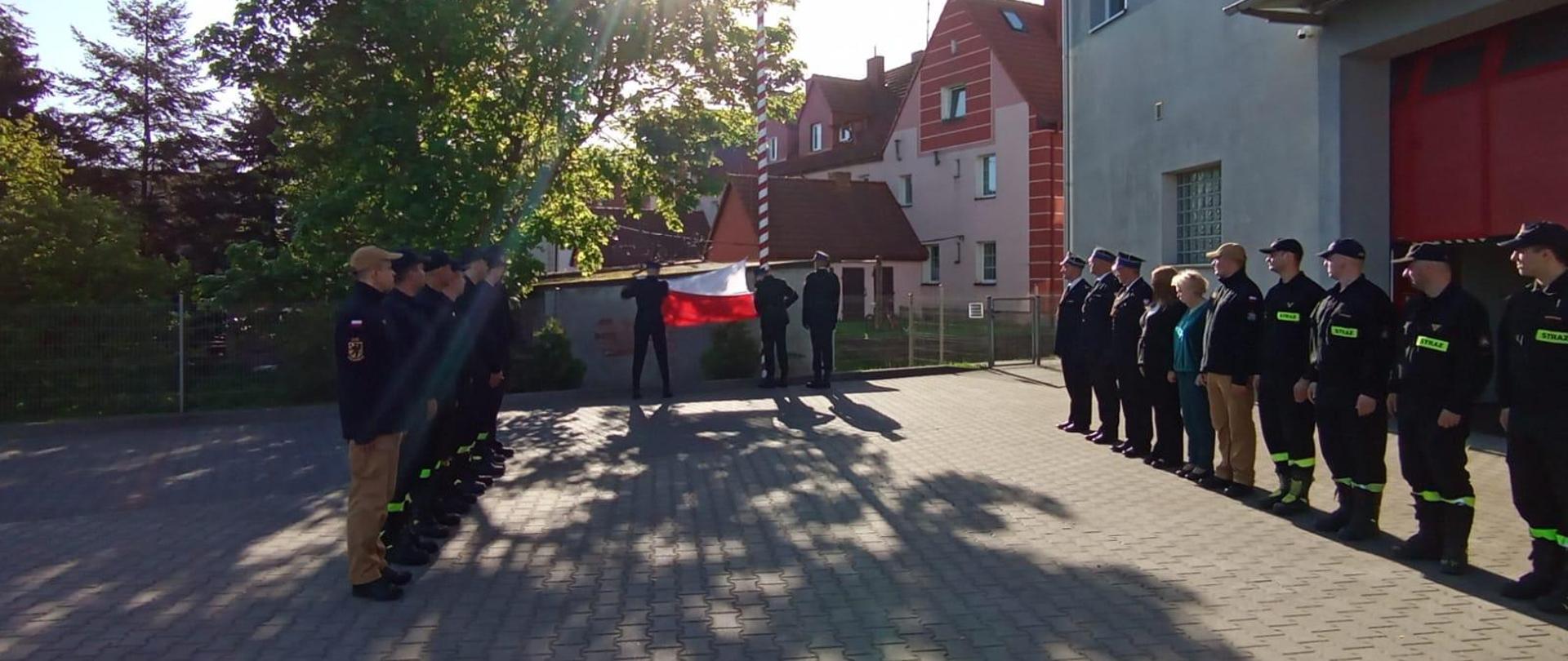 Strażacy oraz pracownicy służby cywilnej Komendy Powiatowej Państwowej Straży Pożarnej w Lęborku podczas uroczystego podniesienia flagi państwowej na maszt. Wszyscy stoją na baczność na placu. Rozłożona flaga trzymana przez strażaka pocztu flagowego w trakcie uroczystego podniesienia na maszt.