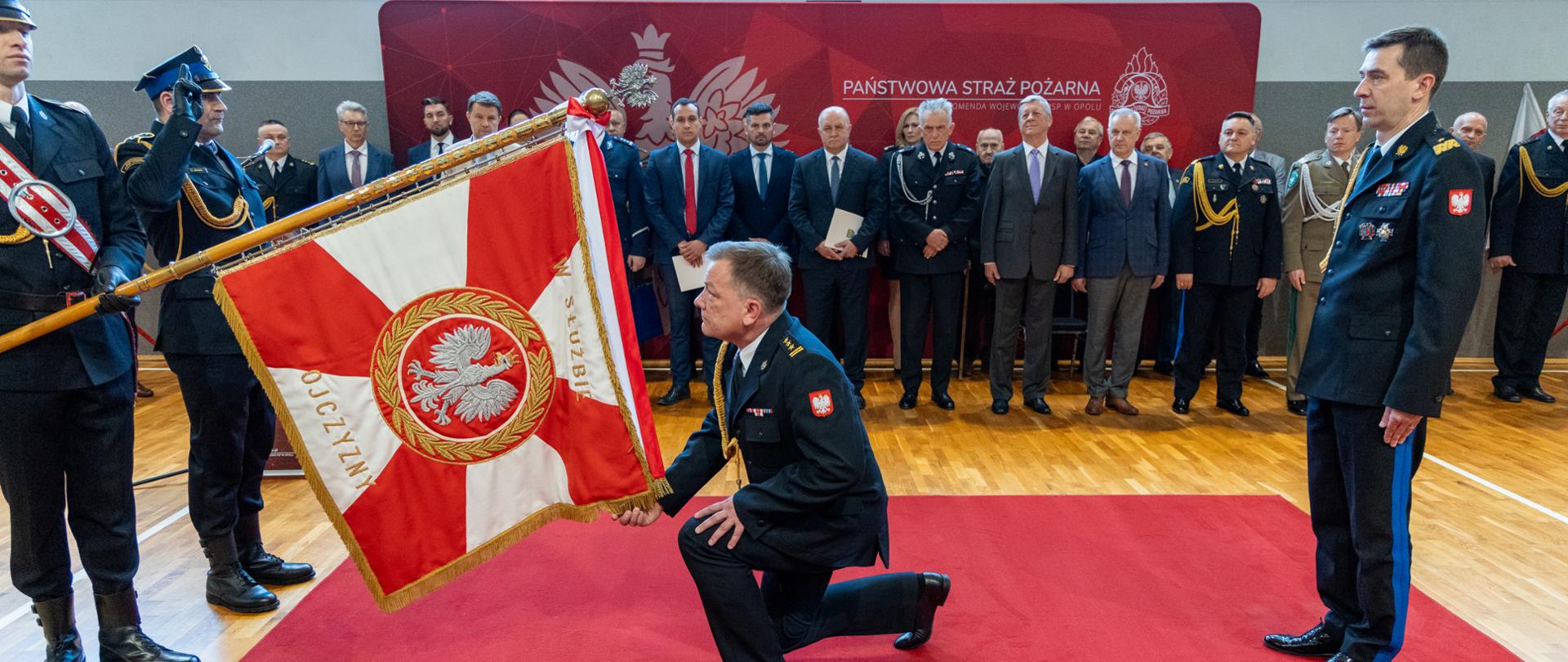 Opolski komendant wojewódzki PSP klęka przed sztandarem oddając mu hołd i ucałować. Na zdjęciu z stoi z prawej strony komendant główny PSP. w tle uczestnicy ceremonii powołania komendanta.