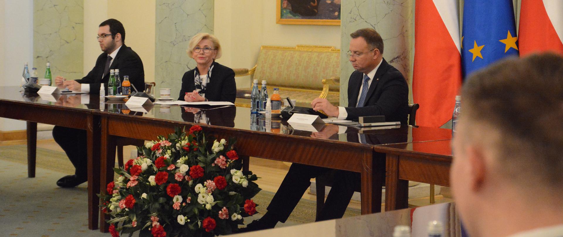 na zdjęciu widoczny z prawej strony Prezydent Duda, z prawej strony siedzi Wiceminister Marzena Machałek. siedzą przy stole, przed nimi biało czerwone kwiaty. z prawej strony prezydenta stoją 2 flagi polski i po środku nich flaga UE