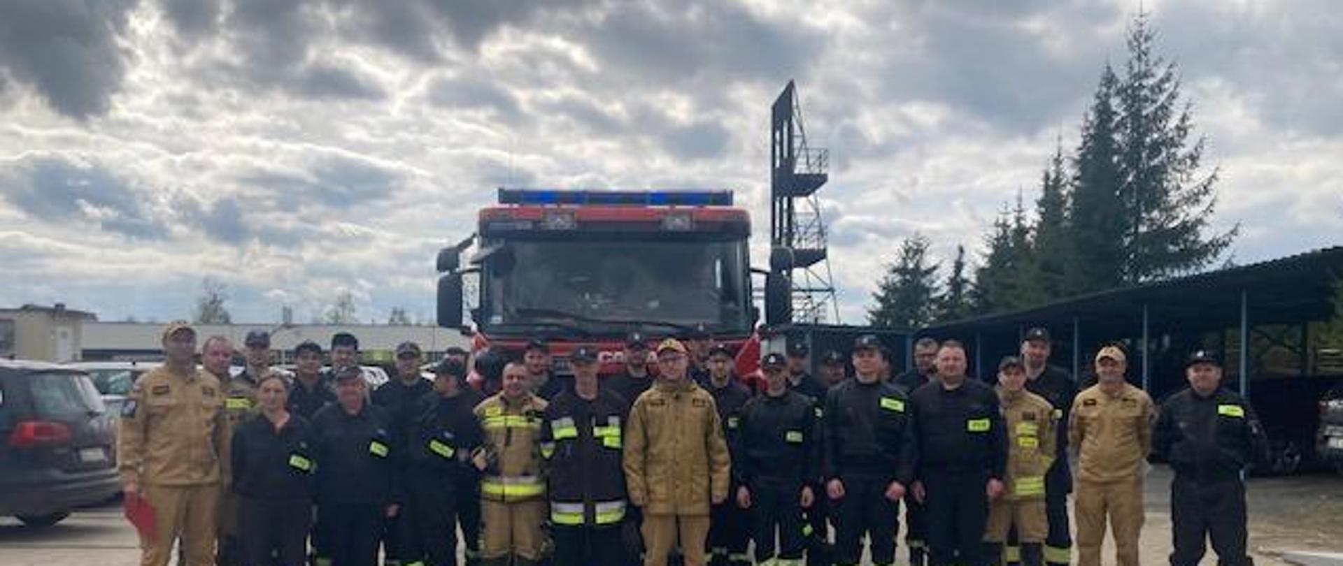 Wspólne zdjęcie dowódców jrg, zastępcy Komendanta Powiatowego w mundurach służbowych, oraz strażaków Ochotniczych straży pożarnych na tle samochodu ratowniczo-gaśniczego.