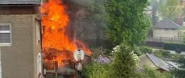 Zdjęcie przedstawia pożar budynku mieszkalnego - pali się drewniana weranda. Płomienie obejmują całą drewnianą werandę budynku.