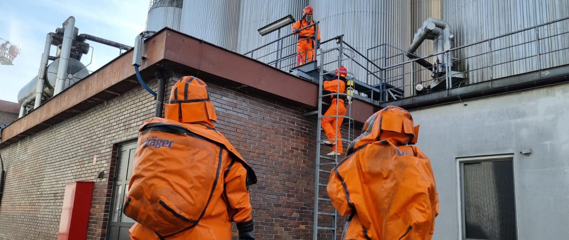 Zdjęcie przedstawia dwóch pracowników zakładu w chemicznych ubraniach gazoszczelnym. Kolejnych dwóch strażaków w ubraniach ochrony chemicznej z założonymi aparatami ochrony dróg oddechowych na plecach wchodzą na dach budynku po drabinie metalowej.