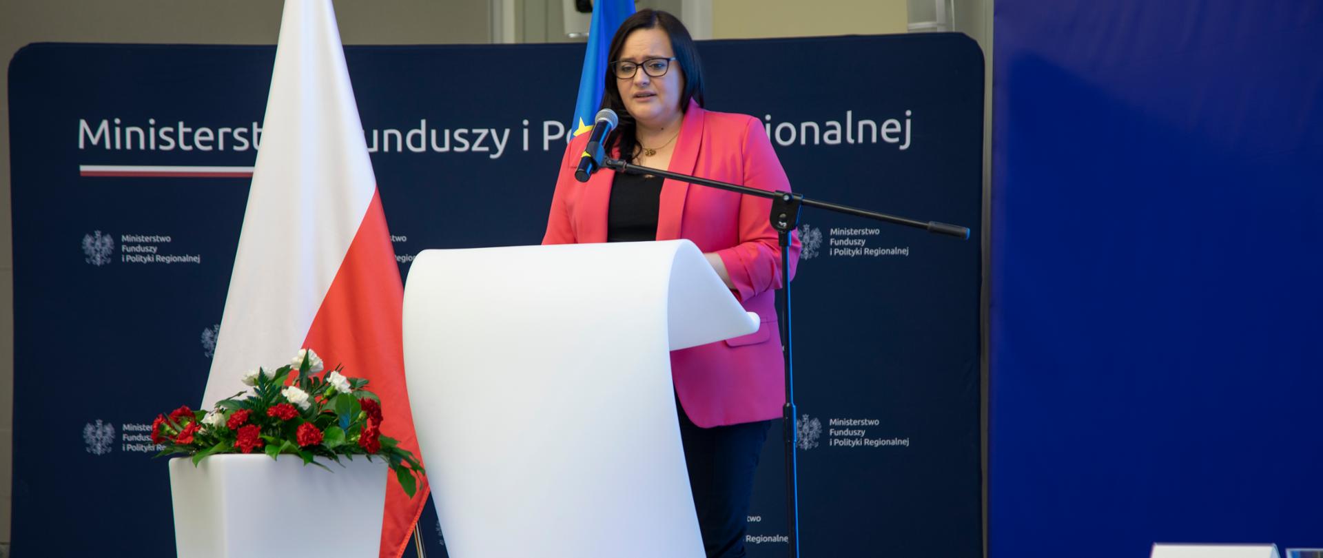 Na zdjęciu stoi przy mównicy wiceminister Małgorzata Jarosińska-Jedynak, w tle flagi Polski i Unii Europejskiej oraz granatowy baner z napisem Ministerstwo Funduszy i Polityki Regionalnej.