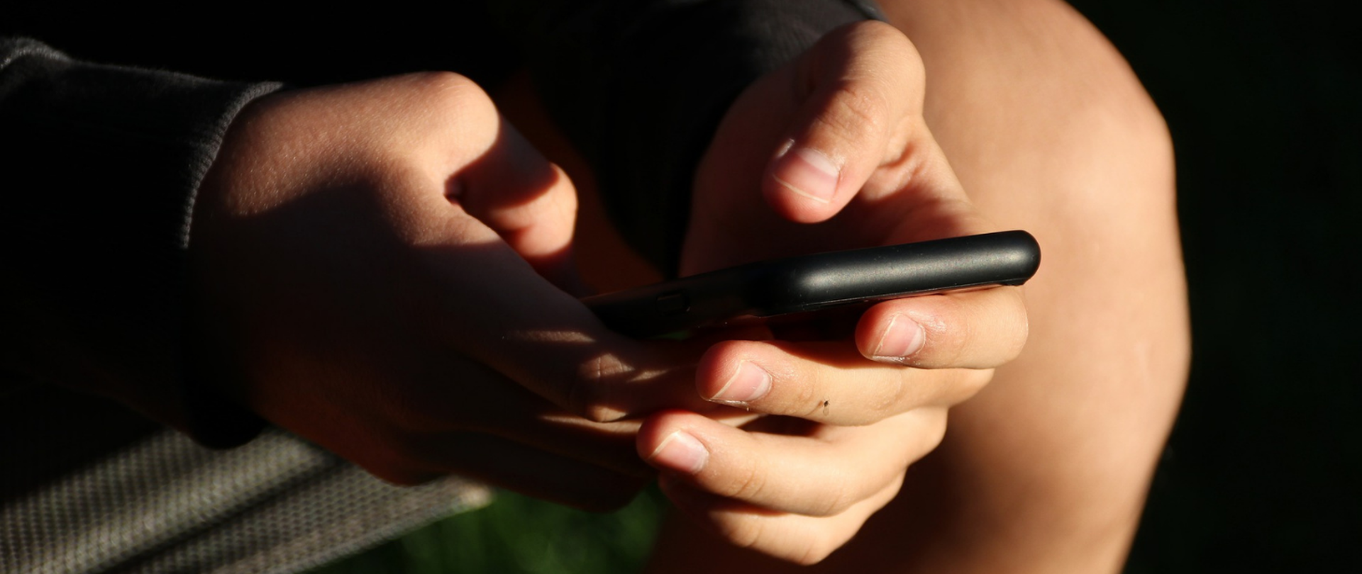 Grafika przedstawia urządzenie mobilne (smartfon) trzymane w dłoniach młodej osoby