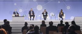 Szczyt Cyfrowy ONZ – IGF 2021. Na scenie, na fotelach siedzi trzech mężczyzn i kobieta. Jednym z nich jest minister Janusz Cieszyński. Z przodu widać kilka rzędów publiczności. Fot. PAP