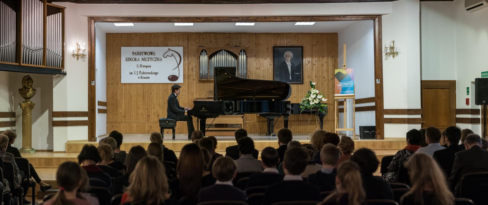 Zdjęcie sali koncertowej PSM w Koninie, pianista siedzi przy fortepianie, na widowni publiczność, na scenie białe kwiaty i plakat konkursu pianistycznego. W tle logo szkoły i portret I. J. Paderewskiego, na ścianie prospekt organów