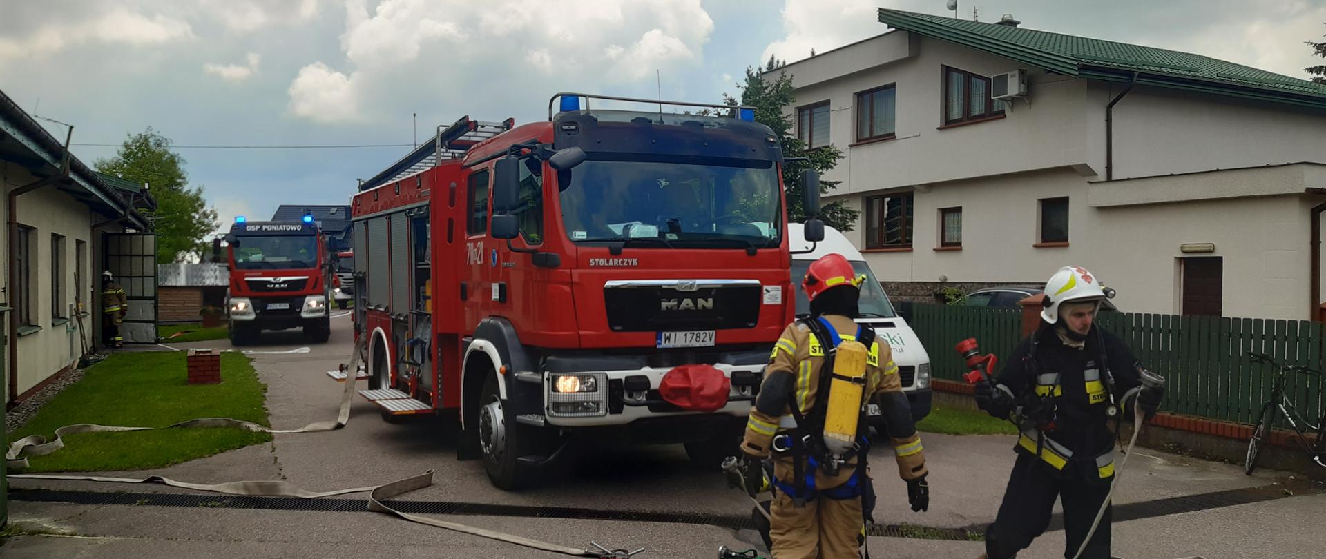 Samochody ratowniczo - gaśnicze straży pożarnej stoją między obiektami stolarni. Strażacy OSP i PSP w ubraniach specjalnych rozkładają węże strażackie. 