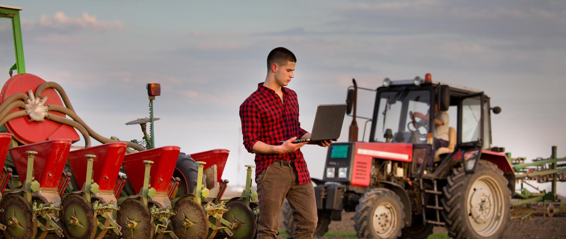 Young farmer with laptop standing in field in front of tractor with smart sowing equipment