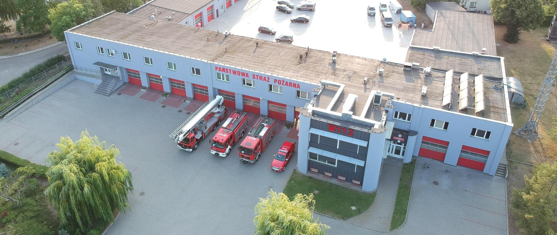 Zdjęcie przedstawia budynek Komendy Miejskiej Państwowej Straży Pożarnej w Koninie. Zdjęcie zrobione z góry. Przed budynkiem na placu stoją samochody strażackie. 