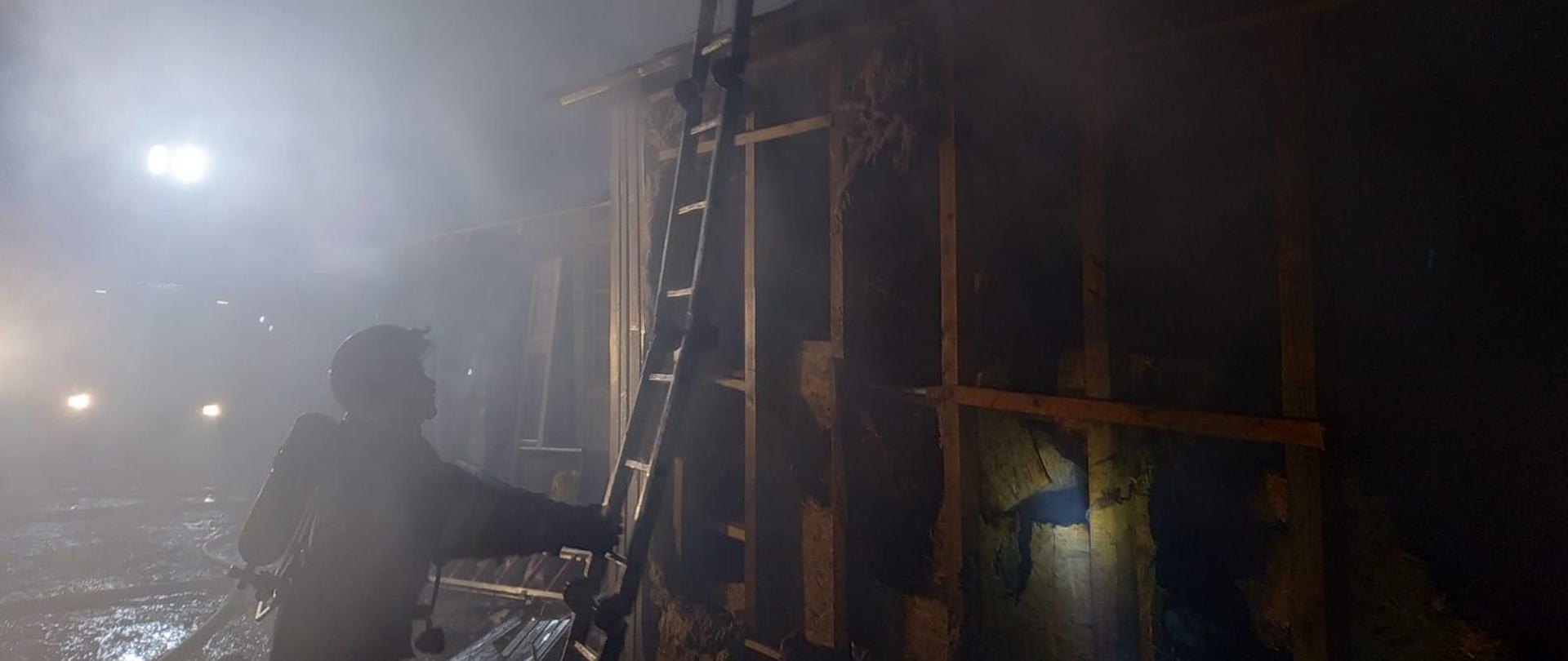 Zdjęcie przedstawia częściowo uszkodzony pożarem budynek stolarni. Ściana budynku jest zdemontowana - widać zniszczoną izolację z wełny. Po środku zdjęcia znajduje się strażak składający drabinę po zakończonej akcji gaśniczej. Zdjęcie wykonane w porze nocnej.