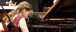 Zdjęcie przedstawiające pianistkę - Magdalenę Dzikoń na scenie filharmonii. W tle orkiestra symfoniczna. 