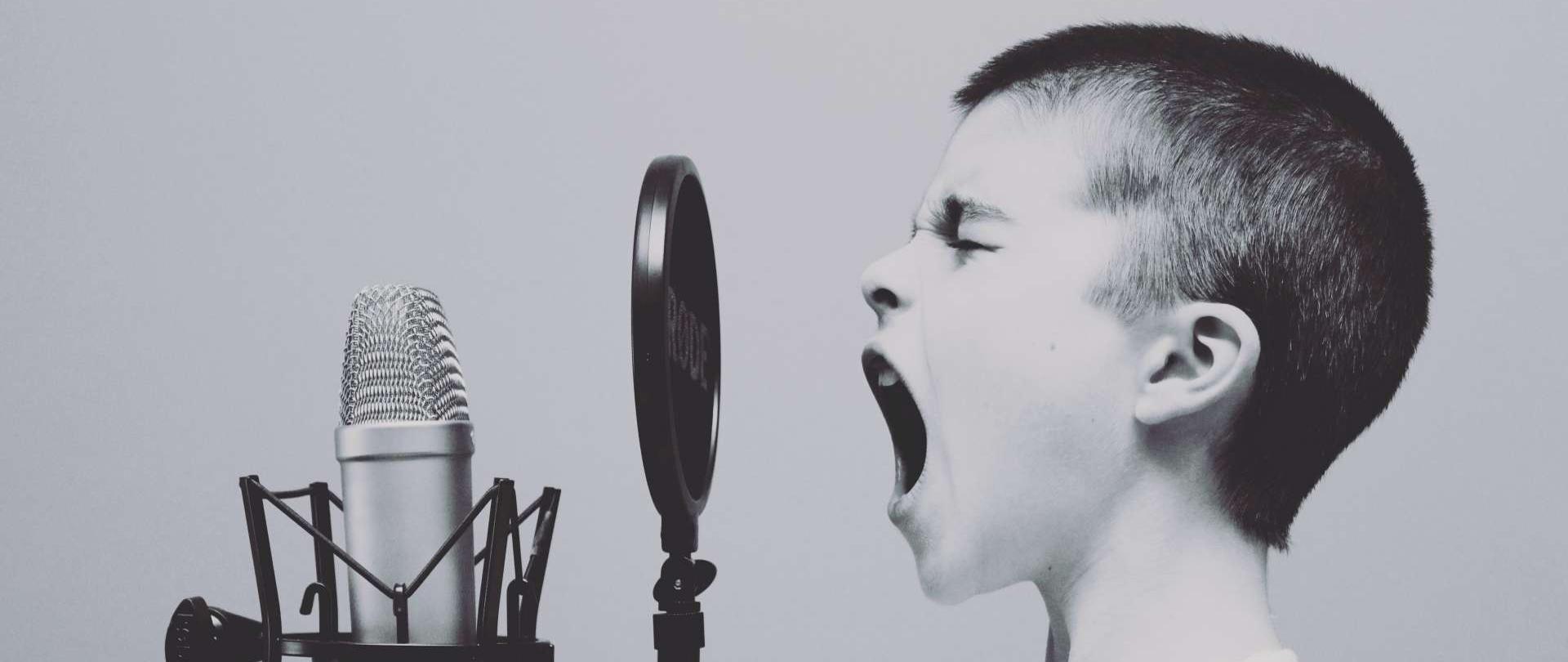 zdjęcie przedstawia ciemnowłosego chłopca ubranego w biały podkoszulek, chłopiec śpiewa do mikrofonu studyjnego