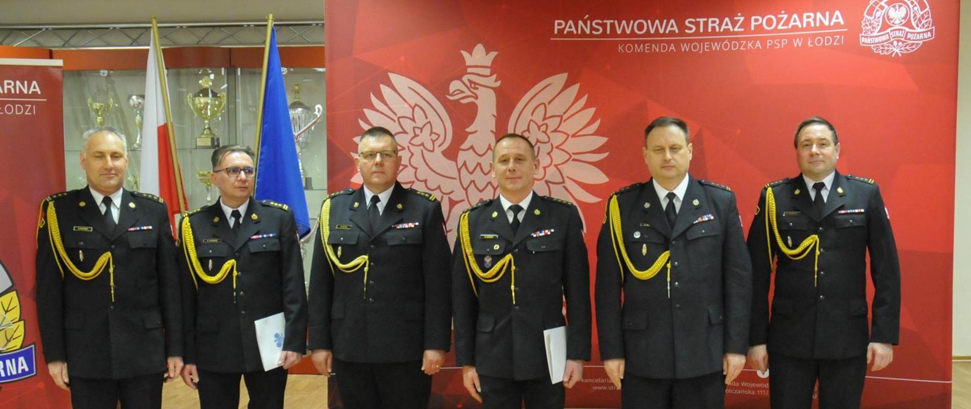 Komendanci PSP stojący w mundurach wyjściowych na czerwonym tle