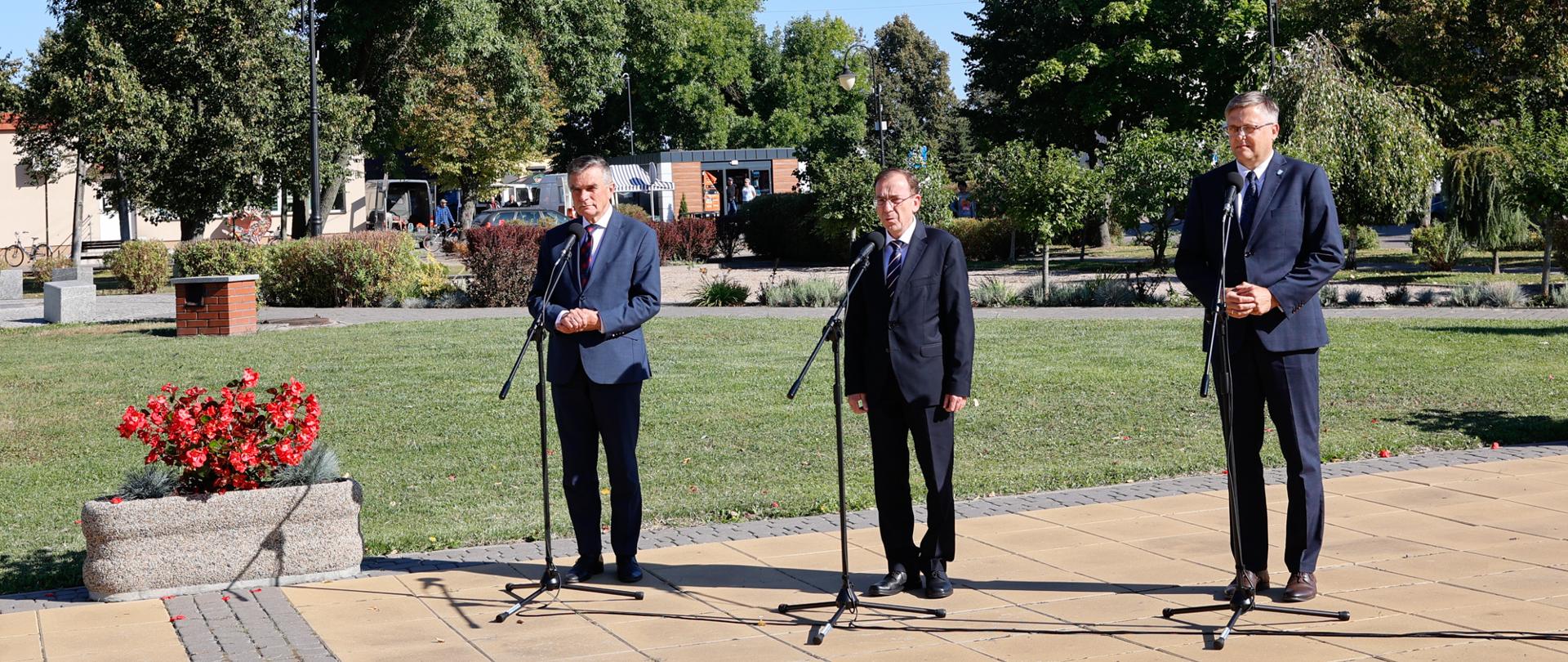 Trzech mężczyzn w garniturach stoi przy mikrofonach.