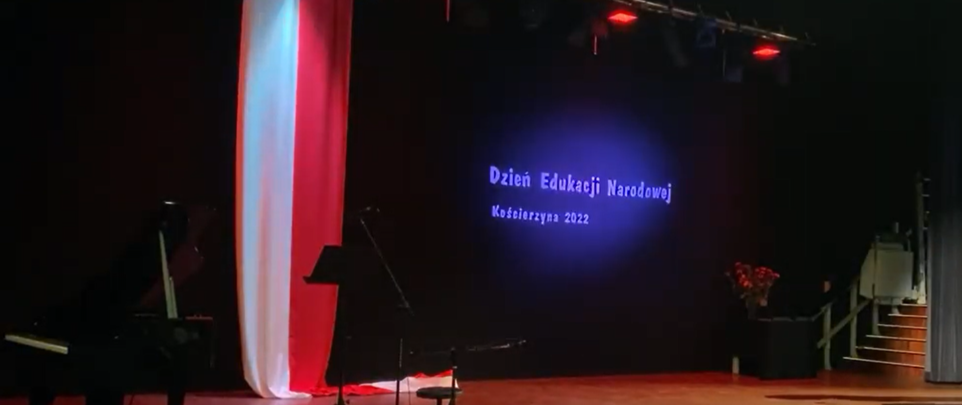 Na ciemnym tle w centralnej części podświetlony na niebiesko napis "Dzień Edukacji Narodowej Kościerzyna 2022". Z lewej strony obrazka w pionie biało- czerwona flaga, a po jej lewej stronie można dostrzec czarny fortepian.