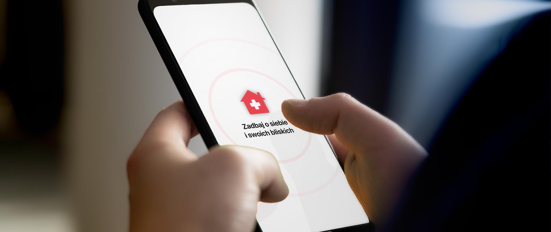 Smartfon w dłoniach - bliskie ujęcie. Na ekranie ikona domku z białym krzyżykiem i napis: Zadbaj o siebie i swoich bliskich