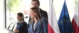 Minister funduszy i polityki regionalnej Katarzyna Pełczyńska-Nałęcz stoi przy mównicy, po jej prawej stronie stoi wiceminister Jan Szyszko i tłumaczka języka migowego, za ich plecami flagi Polski i UE