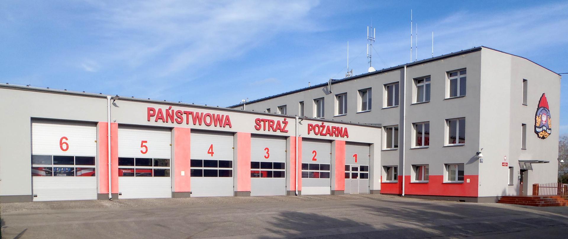 Budynek komendy PSP w Piasecznie. Widoczne bram wyjazdowych i budynek biurowy 3 kondygnacyjny. Nad garażami napis Państwowa Straż Pożarna, nad wejściem do budynku biurowego logo PSP. 