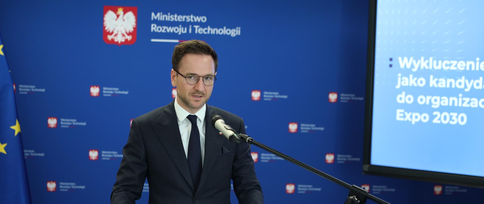 Minister Waldemar Buda stoi przy mównicy i przemawia. W tle ciemnoniebieska ścianka z logo ministerstwa.