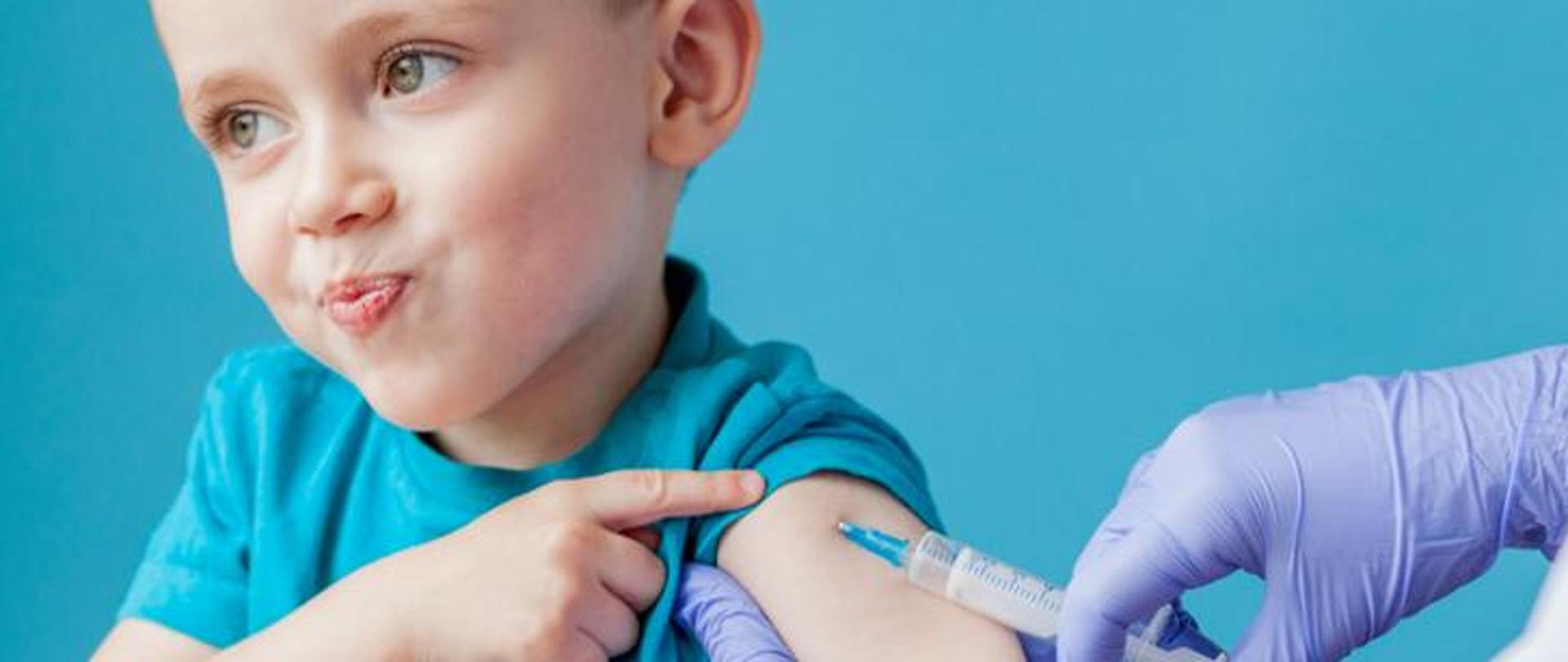 na niebieskim tle dziecko wskazuje ręką przedramię w którym podawana jest szczepionka