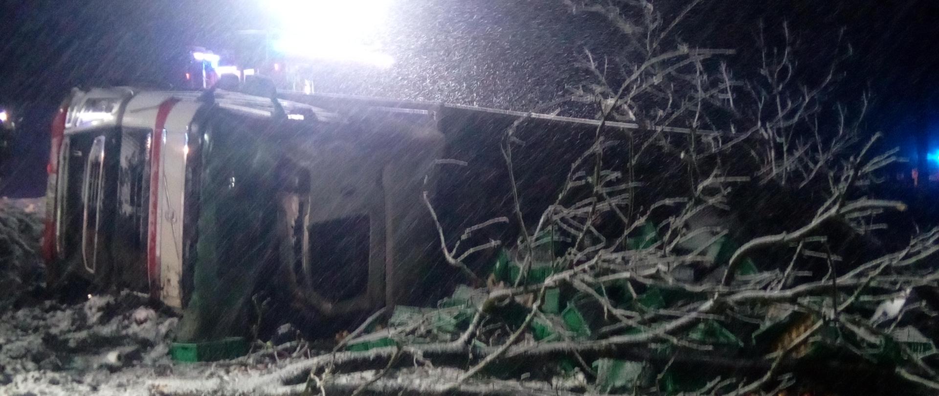 Na zdjęciu widać przewrócony ciągnik siodłowy z naczepą a obok złamane drzewo. 