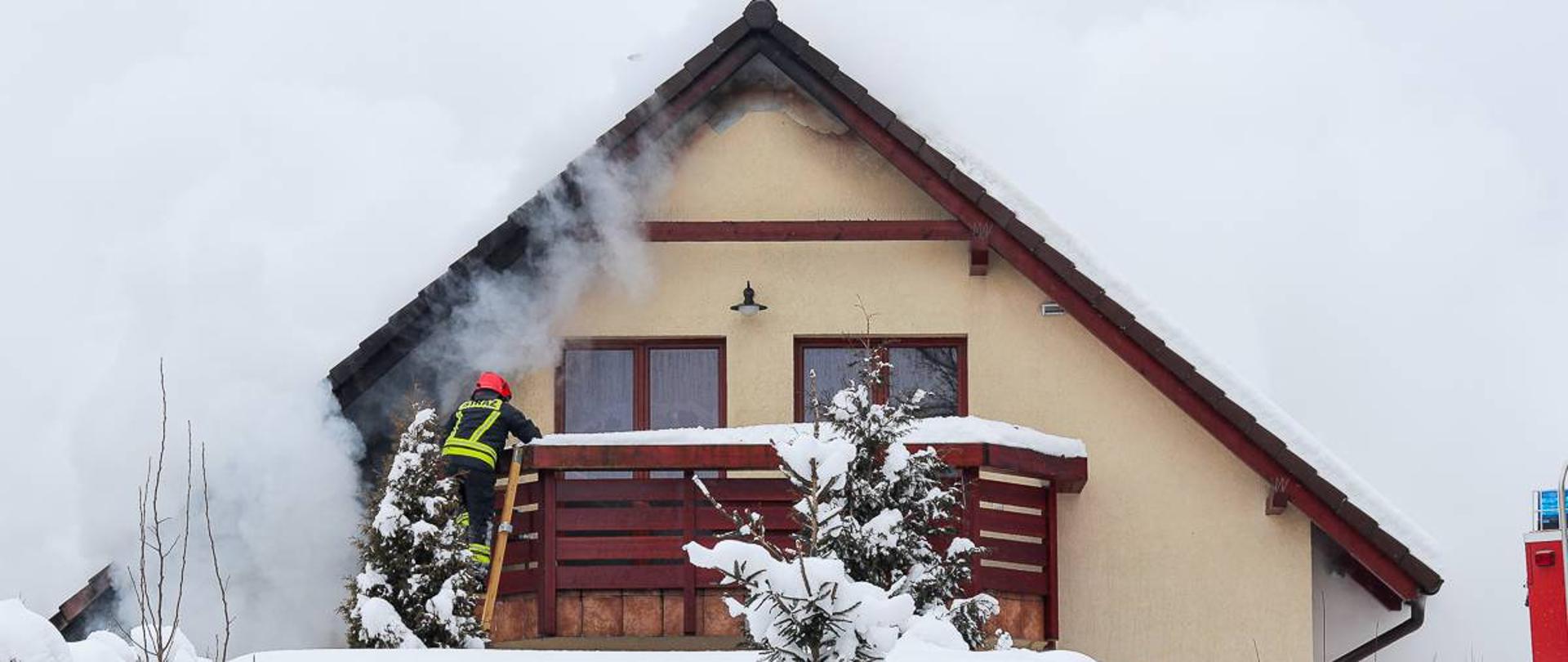 Zdjęcie przedstawia strażaka wchodzącego po drabinie na balkon palącego się budynku. Na zdjęciu widoczny dym i para unosząca się z nad dachem obiektu.