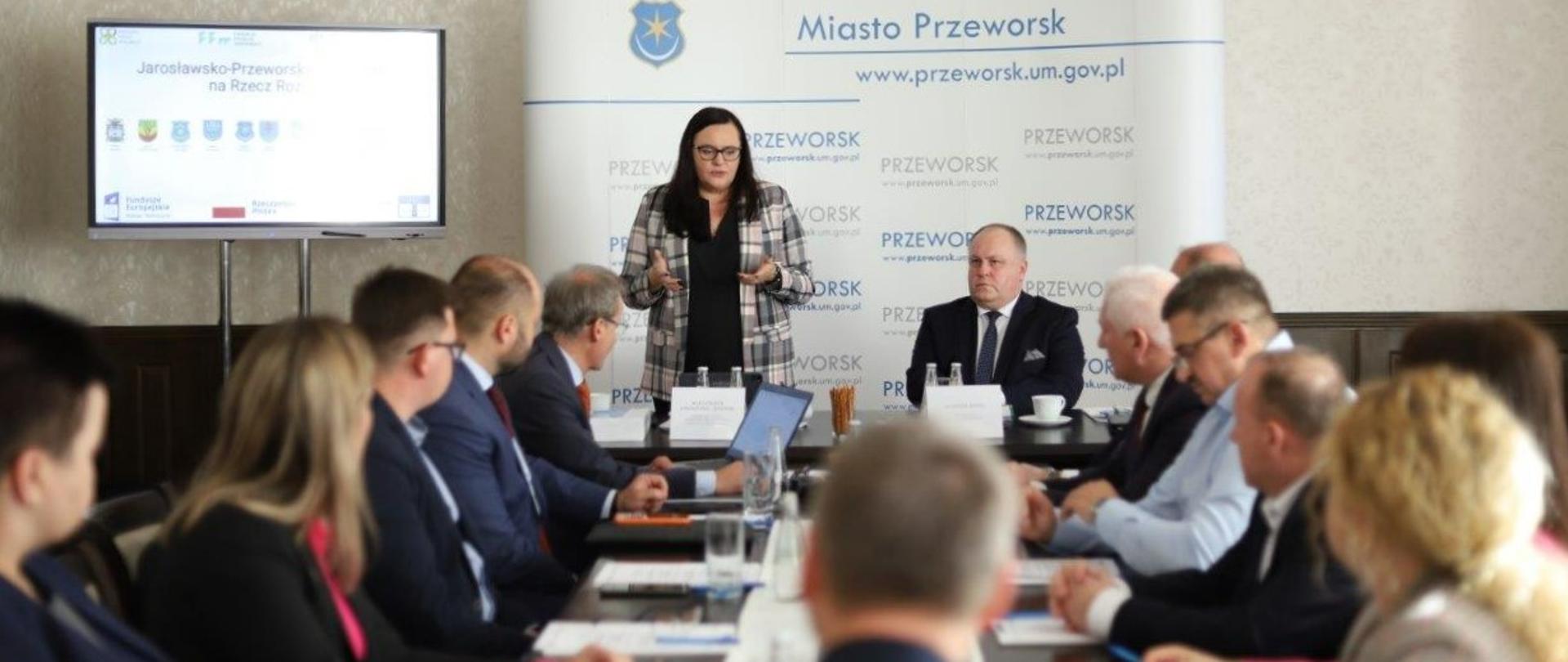 W sali grupa osób siedzi wokół stołu. Na tle ścianki, przy brzegu stołu stoi wiceminister Małgorzata Jarosińska-Jedynak.