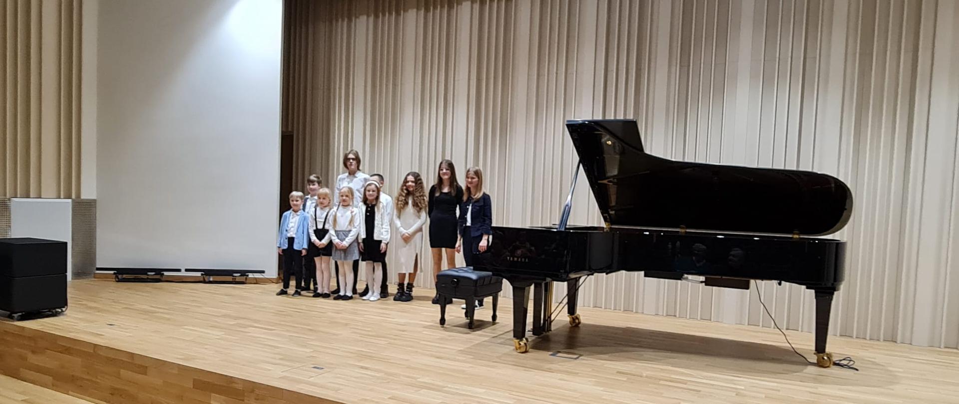 Na zdjęciu grupa odświętnie ubranych uczniów, którzy wystąpili podczas koncertu fortepianowego. Obok grupy dzieci czarny fortepian. Tło obrazka a odcieniach brązu.