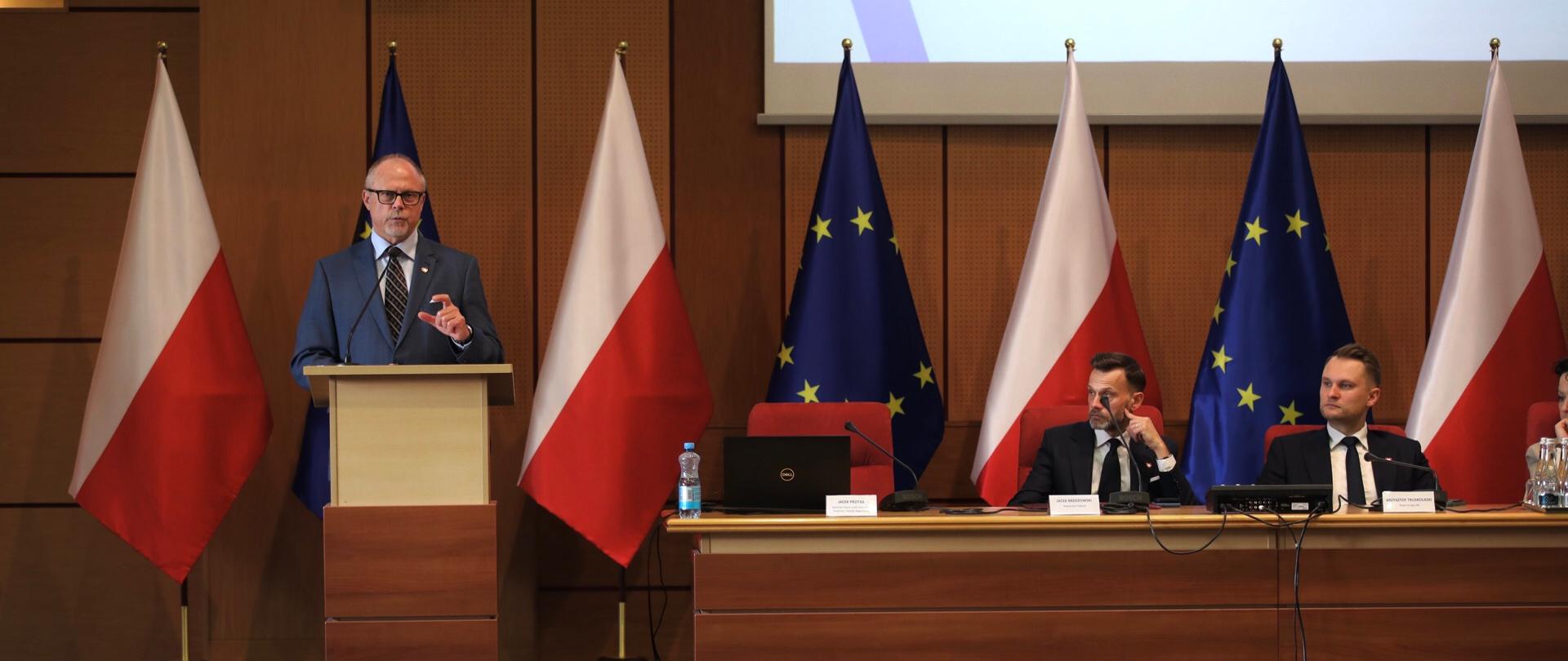 W sali konferencyjnej wiceminister Jacek Protas w mównicy. Obok przy stole prezydialnym siedzi dwóch mężczyzn.