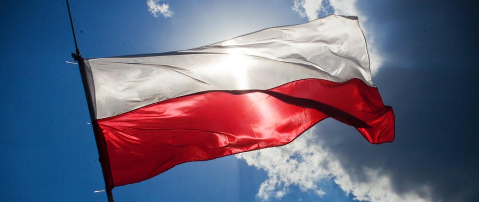 Flaga Polski na tle błękitnego nieba z chmurami przez które przebijają promienie słoneczne.