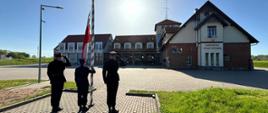 Zdjęcie przedstawia strażaków Komendy Powiatowej Państwowej Straży Pożarnej w Gołdapi wchodzących w skład pocztu flagowego, podczas podnoszenia flagi Rzeczypospolitej Polskiej na maszt przy akompaniamencie hymnu państwowego z okazji Dnia Flagi Państwowej Rzeczypospolitej Polskiej. Na zdjęciu w oddali widoczni są również strażacy Komendy Powiatowej Państwowej Straży Pożarnej w Gołdapi na tle pojazdów pożarniczych zlokalizowanych w budynku strażnicy