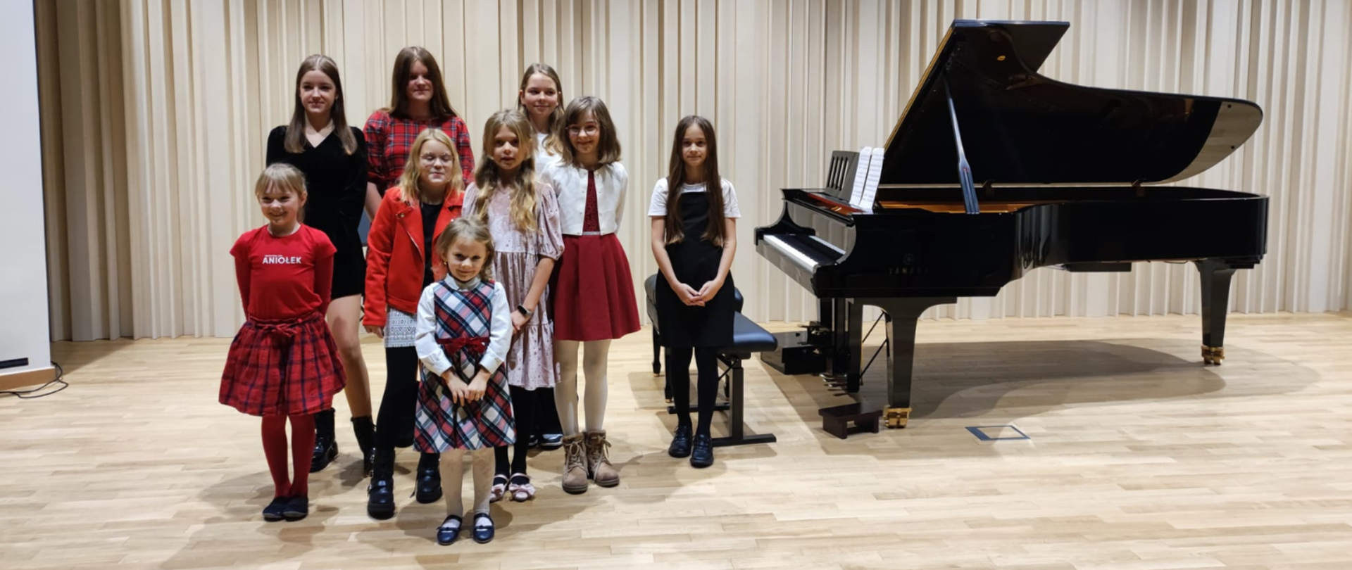 Na zdjęciu grupa uczniów- same dziewczynki w różnym wieku. Obok nich po prawej stronie stoi czarny fortepian.