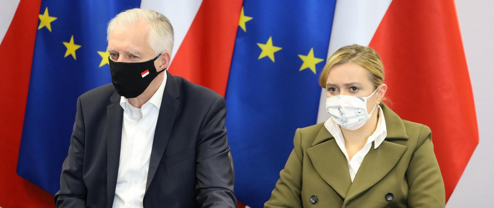 Jarosław Gowin i Olga Semeniuk siedzący za biurkiem. Za nimi flagi polska i unijna.