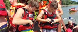 Obóz szkoleniowo-wypoczynkowy MDP w Brennie. Na plaży odbywają się zajęcia z zakresu ratownictwa wodnego oraz bezpiecznego wypoczynku nad wodą. Obozowicze przygotowuj się do rejsu patrolowego po Jeziorze Białym. Dwóch z nich ubiera kapoki. Z prawej strony pontony ratownicze straży pożarnej .