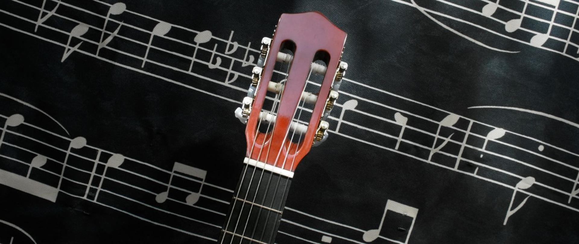 zdjęcie - gryf gitary klasycznej, sześciostrunowej, na tle czarnej tablicy z pięciolinią i nutami