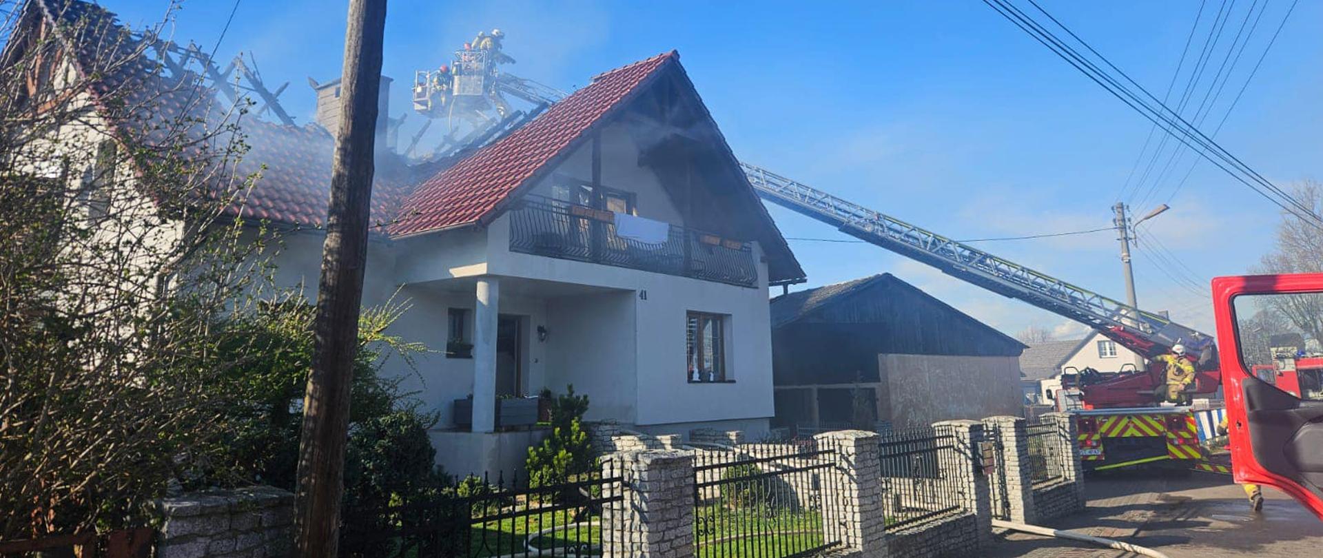 Na zdjęciu znajduje się drabina hydrauliczna , z której działania prowadzą strażacy w SOUO, na pierwszym planie znajduje się budynek mieszkalny z nadpalonym dachem