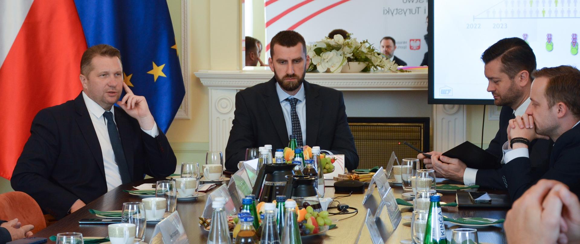 Uczestnicy spotkania siedzą i rozmawiają przy stole, za nimi flagi Polski i UE oraz duży ekran.