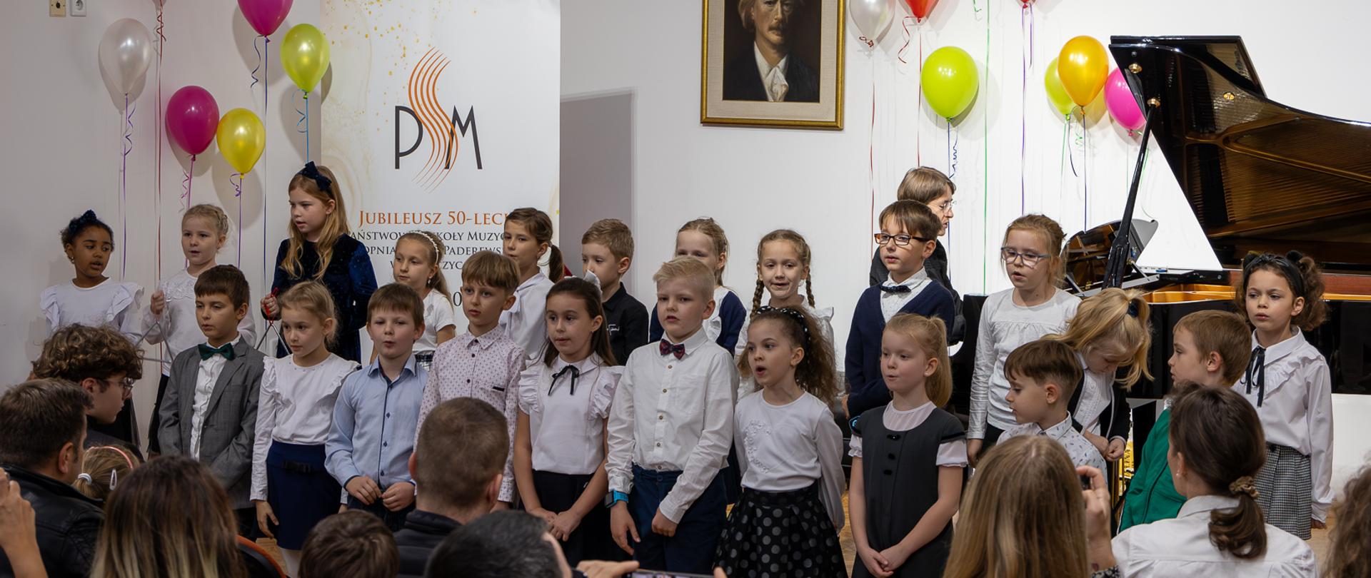 Pierwszoklasiści śpiewają wspólnie na scenie PSM w Głubczycach podczas uroczystości pasowania na ucznia. Przy fortepianie pani Małgorzata Meyse. W dolnej części zdjęcia widoczna publiczność.