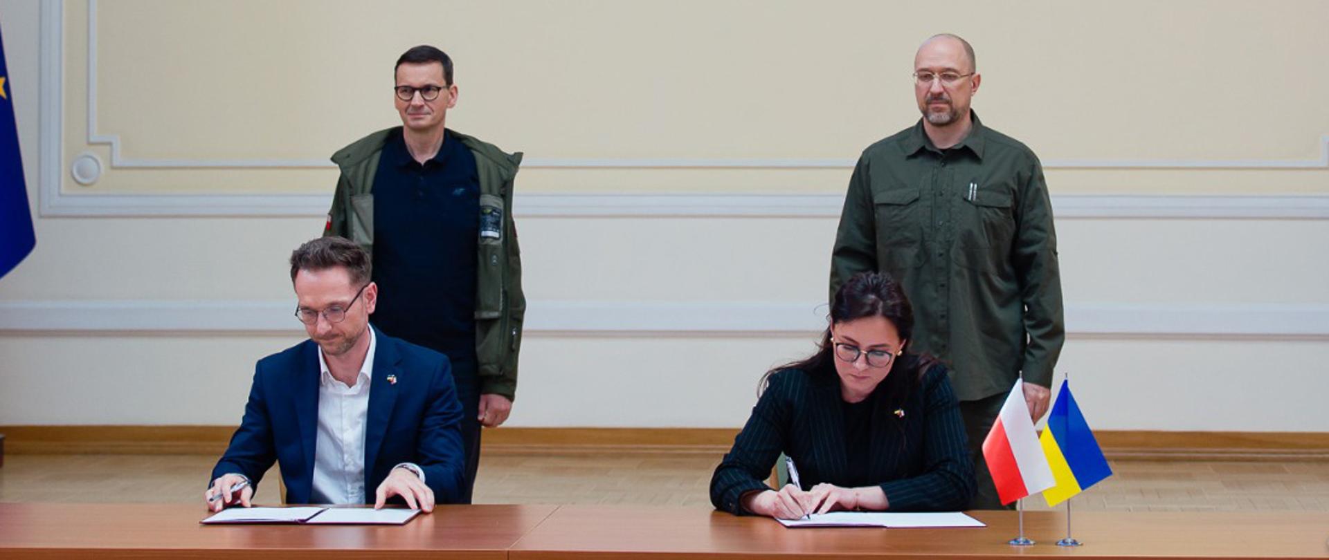 Waldemar Buda i Julia Svyrydenko siedzą przy stole, podpisują porozumienie. Za nimi stoja premierzy obu krajów.