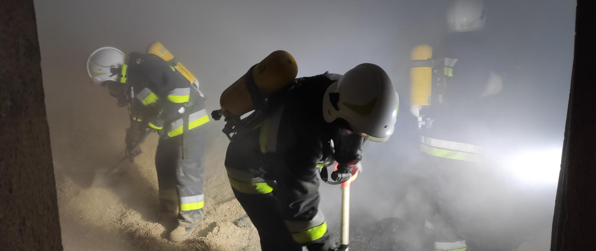 Na zdjęciu strażacy w ubraniach specjalnych, białych hełmach oraz aparatach oddechowych wyrzucają łopatami trociny. Pomieszczenie jest zadymione.