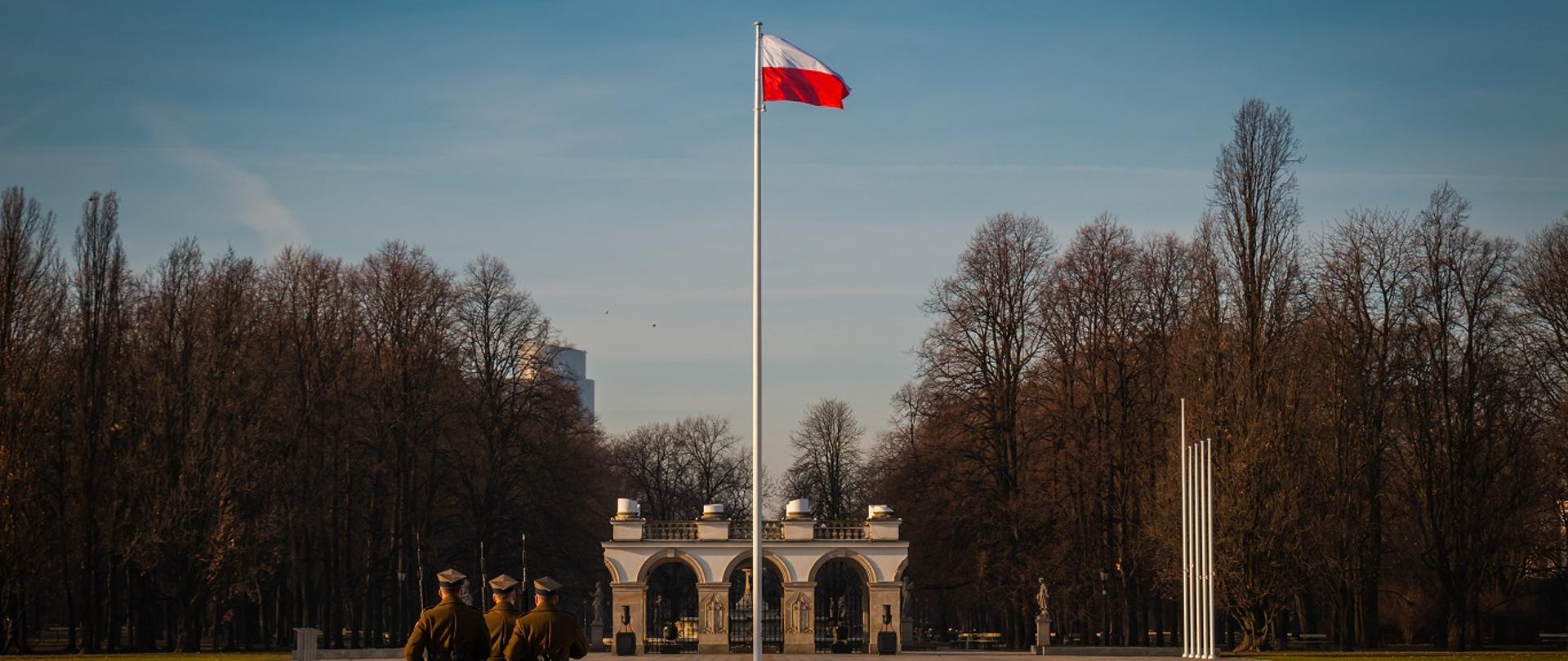 trzech polskich żołnierzy maszerujących po placu, na którym stoi maszt z zawieszoną biało-czerwoną flagą