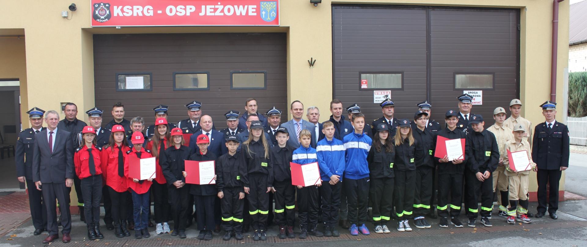 członkowie młodzieżowych drużyn pożarniczych z promesami strażacy oraz goście na tle remizy OSP