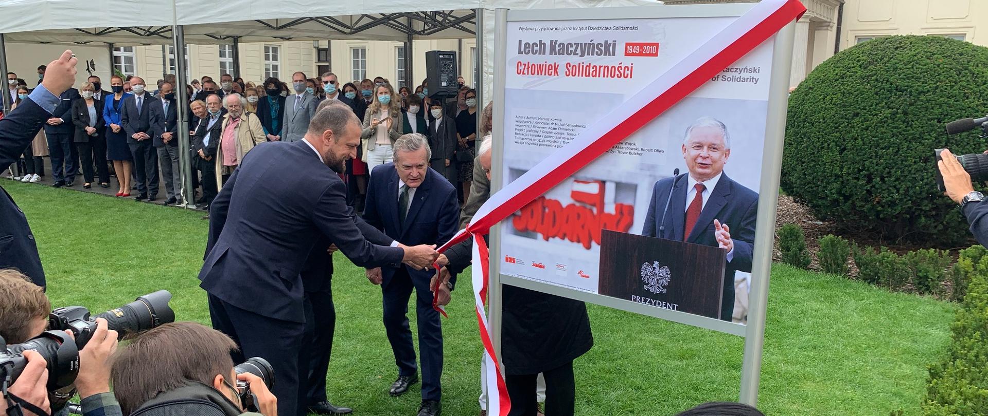 Ludzie w garniturach stoją przed tablicą ze zdjęciem Lecha Kaczyńskiego, za nimi budynek MKiDN.