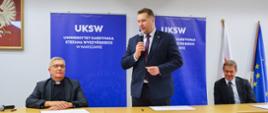 Minister Czarnek stoi za stołem i mówi do mikrofonu, obok niego siedzi dwóch mężczyzn w garniturach, za nimi na białej ścianie niebieskie banery z napisami UKSW.