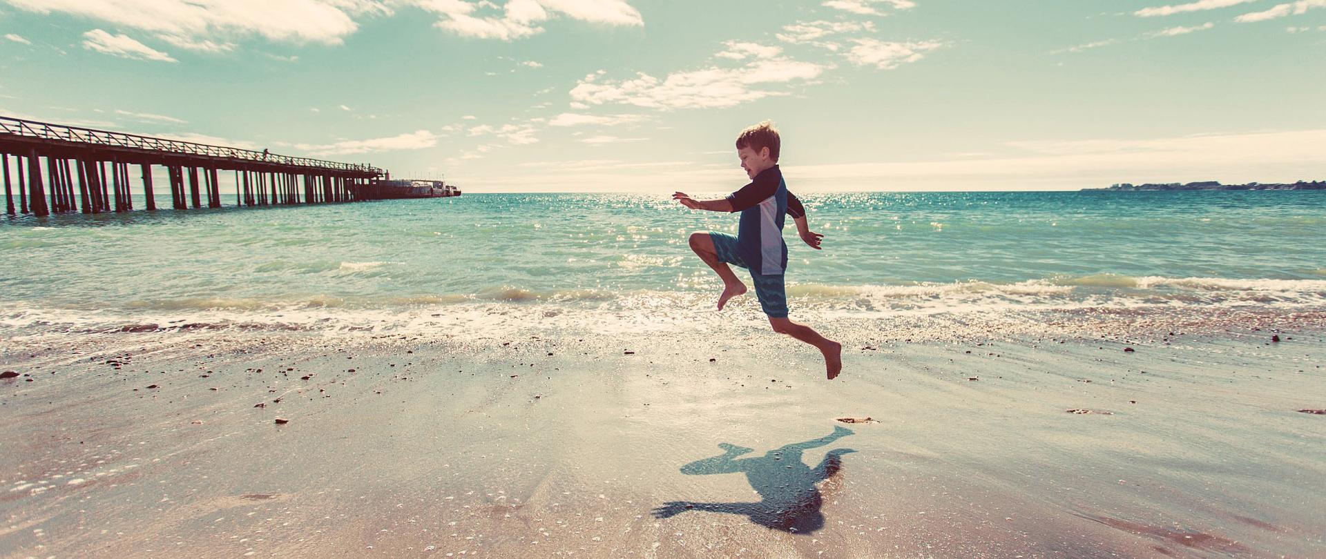 Chłopiec biegnący po plaży na tle morza. Po lewej molo biegnące w głąb morza z zacumowanymi na końcu statkami.